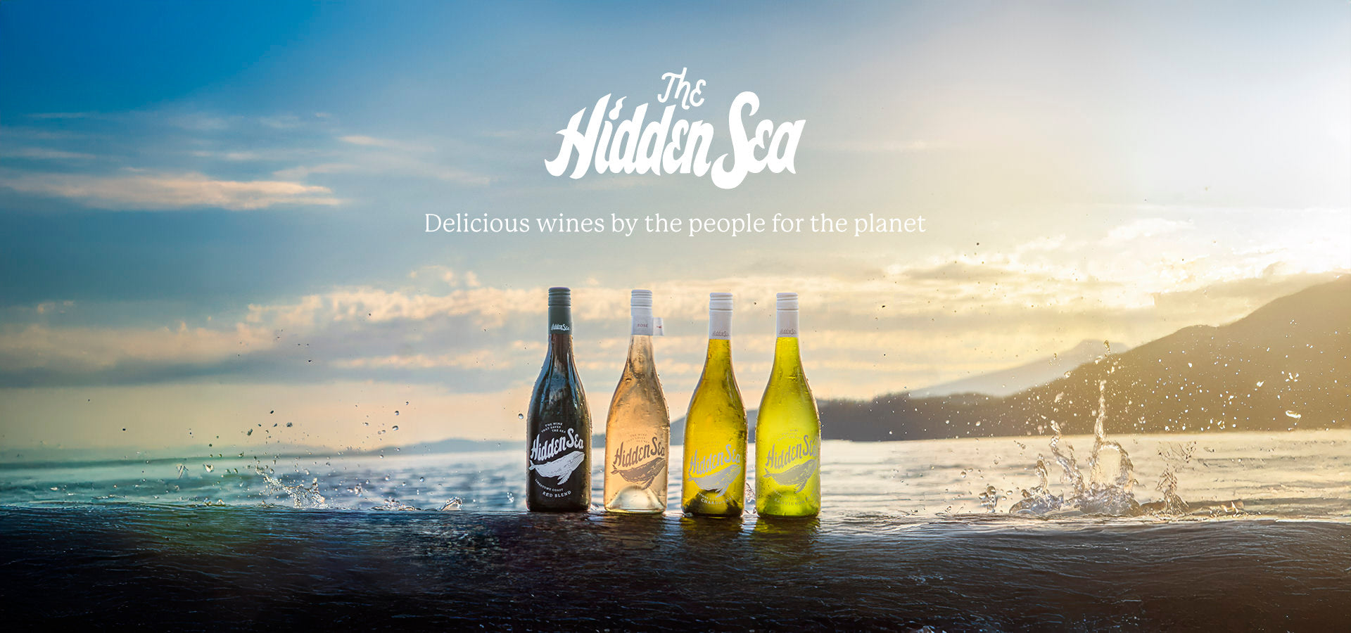 A lineup of four The Hidden Sea wine bottles stands in a row on the ocean's surface, splashed by waves at sunset. The bottles range from dark reds on the left to white and rosé on the right. The sky glows with soft pastel hues and clouds, with mountains visible in the background. Above the bottles, the logo reads “The Hidden Sea” and the tagline says, “Delicious wines by the people for the planet.”